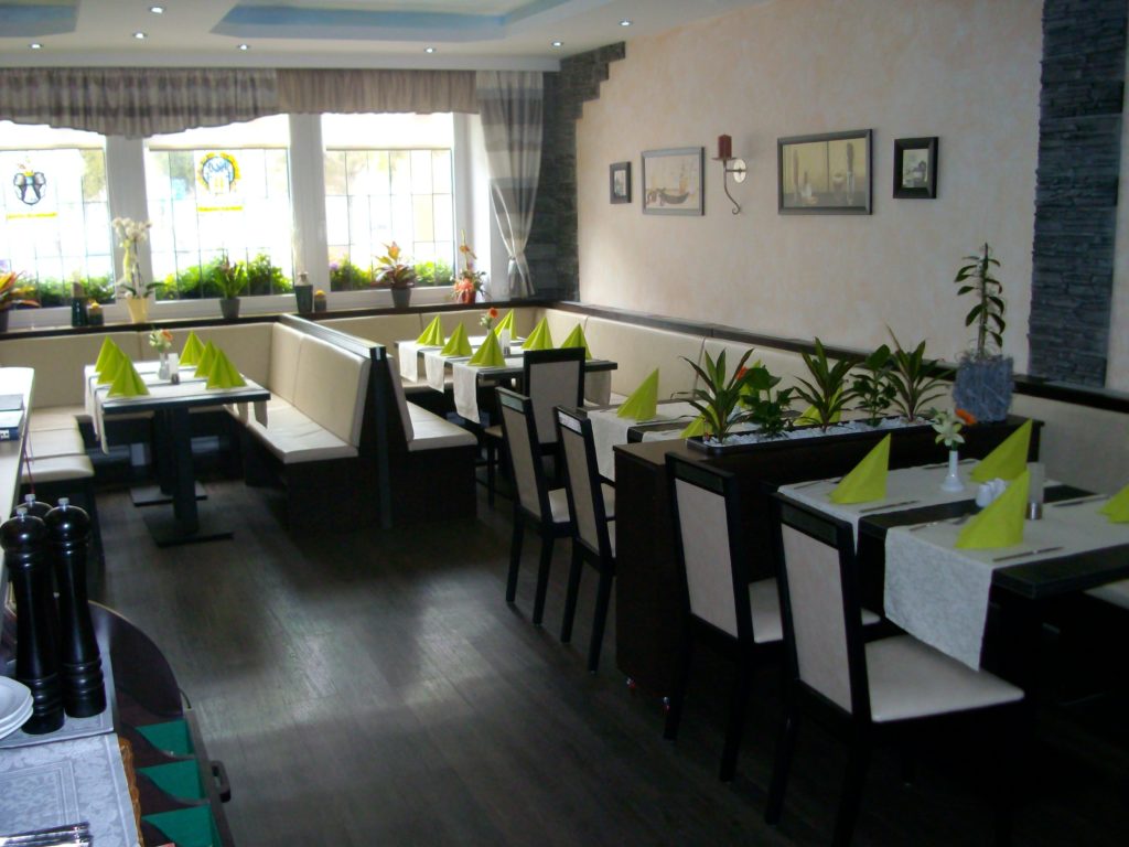 Ein moderner Speiseraum eines Balkan-Restaurants mit Tischen und Stühlen, ordentlich gefalteten grünen Servietten, Zierpflanzen, Wanddekorationen und großen Fenstern, durch die Tageslicht hereinströmt. Der Raum wirkt sauber und einladend.