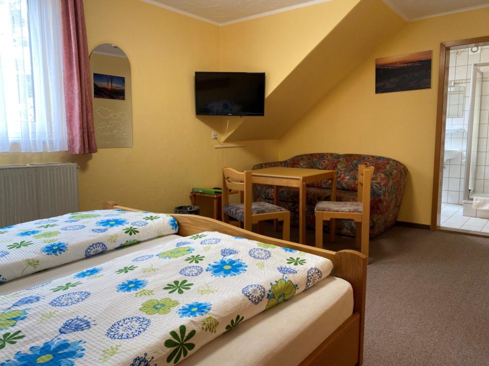 Gemütliches Schlafzimmer mit zwei Einzelbetten, Bettwäsche mit Blumenmuster, einer kleinen Sitzecke mit Tisch und Stühlen, einem an der Wand montierten Fernseher und Blick auf ein gefliestes Badezimmer. Durch ein mit Vorhängen versehenes Fenster fällt Sonnenlicht herein.