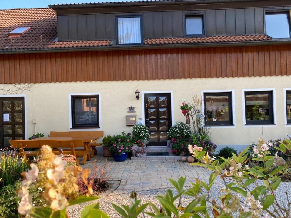 Ein Haus mit einer beigen Fassade, fünf quadratischen Fenstern, einer braunen Tür und Blumentöpfen am Eingang. Links steht eine Holzbank, im Vordergrund stehen Pflanzen, und ein Kopfsteinpflasterweg führt vorbei.