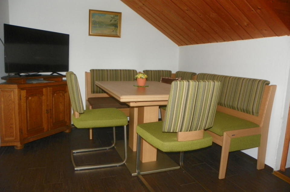 Sitzecke mit zwei Stühlen und einer Eckbank in den Farben grün unter einer Dachschräge mit Holzdecke. Zu sehen ist außerdem eine Kommode mit Fernseher.