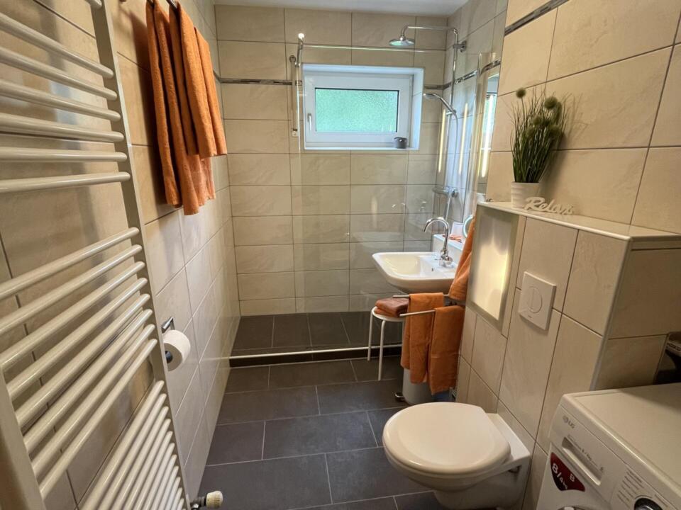 Modernes Badezimmer mit beigen Fliesen, einem wandhängenden WC, einem weißen Waschbecken, einer begehbaren Dusche, einem Handtuchhalter, einer Waschmaschine, orangefarbenen Handtüchern und einem kleinen Fenster. Auf einem Regal über dem WC stehen eine grüne Pflanze und ein Schild mit der Aufschrift „Relax“.