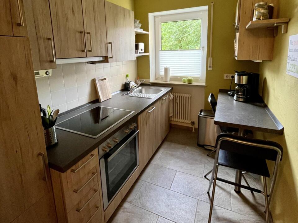 Eine kompakte Küche mit Holzschränken, einem Elektroherd, einer Spüle, einer Kaffeemaschine, einem Fenster und einem kleinen Stehtisch mit einem schwarzen Stuhl. Der Raum hat gelbgrüne Wände und einen Fliesenboden.