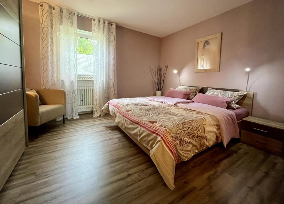 Ein gemütliches Schlafzimmer mit einem Doppelbett, gemusterter Bettwäsche, zwei Nachttischen, sanfter Beleuchtung, einem hellbraunen Sessel, transparenten Vorhängen am Fenster und einem gerahmten Bild an einer taupefarbenen Wand. Der Holzboden sorgt für eine warme Atmosphäre.