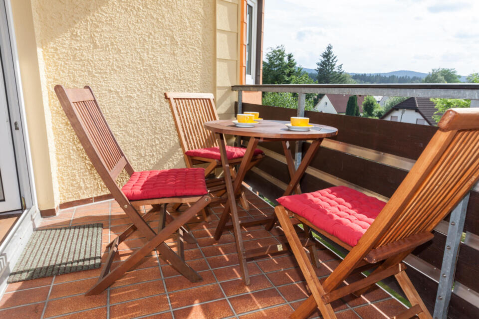 Ein kleiner Balkon mit einem runden Holztisch, drei Holzstühlen mit roten Kissen, zwei gelben Kaffeetassen und einem Blick auf Bäume und Dächer im Hintergrund.