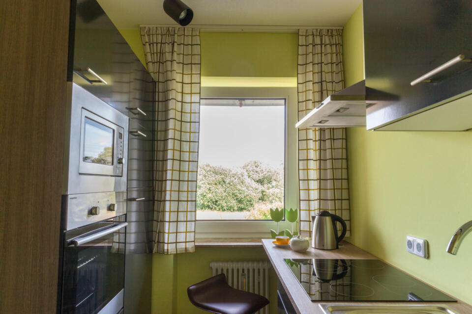 Kompakte, moderne Küche mit grünen Wänden, einem Induktionskochfeld, einem Wasserkocher, einem Backofen, einer Mikrowelle und einem Fenster mit karierten Vorhängen, durch das man auf die Bäume draußen blickt. Ein brauner Barhocker steht an der Theke unter dem Fenster.