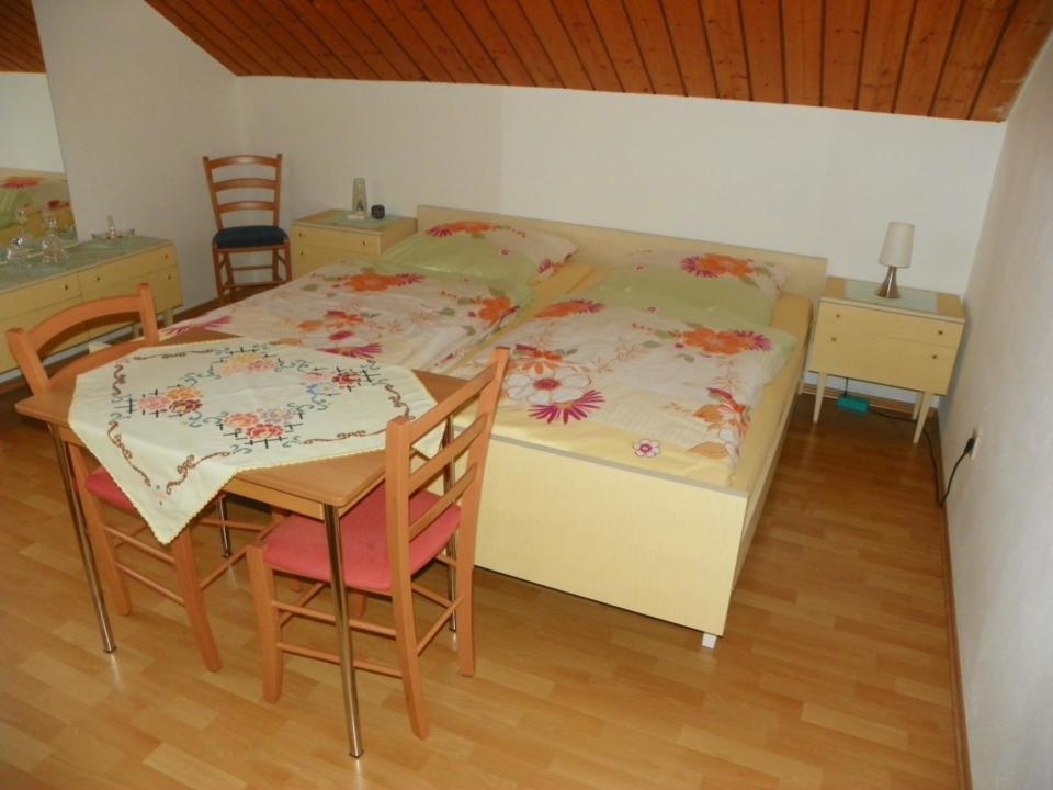 Zu sehen ist die Einrichtung der Ferienwohnung Eitner. Ein Doppelbett mit Bettwäsche in den Farben weiß, orange und pink und davor ein Tisch mit zwei Stühlen.