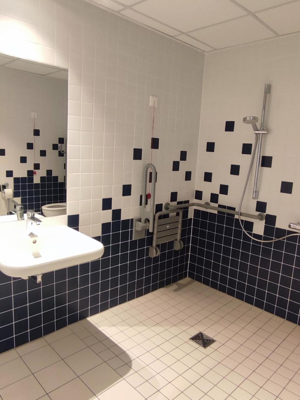 Barrierefreies Badezimmer im Schullandheim Weißenstadt mit einem wandmontierten Waschbecken, Haltegriffen, einer Handbrause, einem Duschsitz und einer roten Notrufschnur. Die Wände sind mit Fliesen in einem ansprechenden weiß-dunkelblauen Karomuster verkleidet.