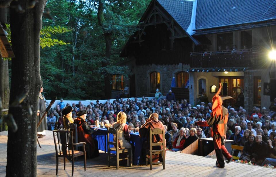 Bei einer Freilichtaufführung im Rahmen der Waldstein-Festspiele sitzen Schauspieler in mittelalterlichen Kostümen an einem langen Tisch, darunter eine Person, die als Hofnarr verkleidet ist. Vor einem großen, rustikalen, von Bäumen umgebenen Gebäude sitzt ein aufmerksamem Publikum.
