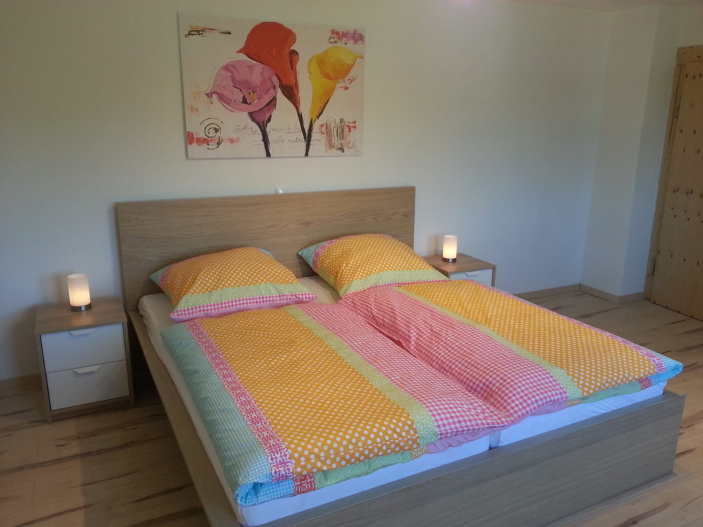 Ein ordentlich gemachtes Bett mit bunter Bettwäsche, flankiert von zwei kleinen Nachttischen mit brennenden Lampen, und ein farbenfrohes Blumenbild, das über dem hölzernen Kopfteil in einem minimalistisch eingerichteten Schlafzimmer hängt.