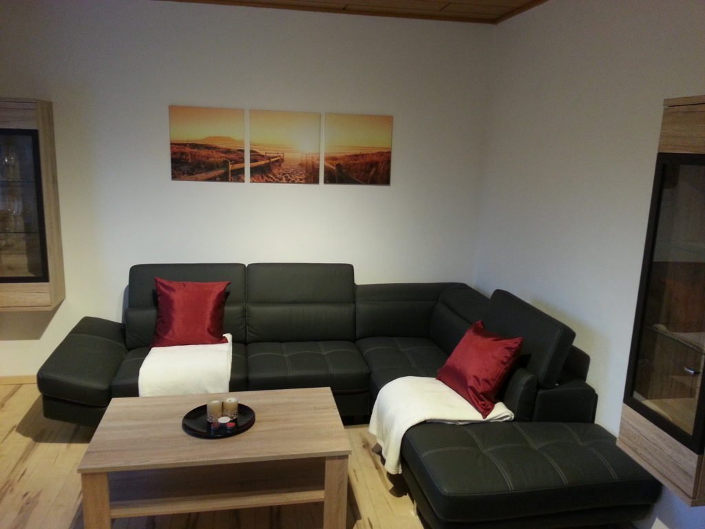 Ein modernes Wohnzimmer mit einem schwarzen Ecksofa, roten und weißen Kissen, einem Couchtisch aus Holz und einem dreiteiligen Kunstdruck mit einem Sonnenuntergang an der Wand. Der Raum verfügt über Holzböden und Wände in neutralen Farbtönen.