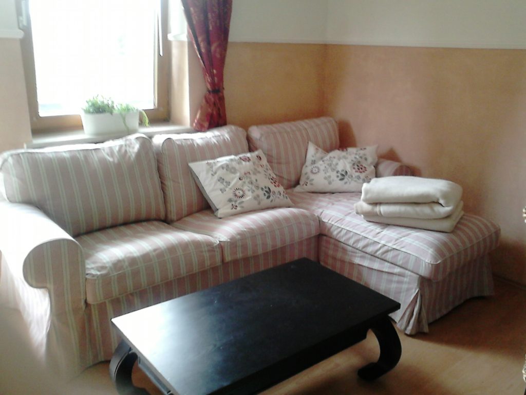 Ein gestreiftes, L-förmiges Sofa mit Blumenkissen und gefalteten Decken steht an einem Fenster mit einer Pflanze und roten Vorhängen; davor steht ein schwarzer Couchtisch.