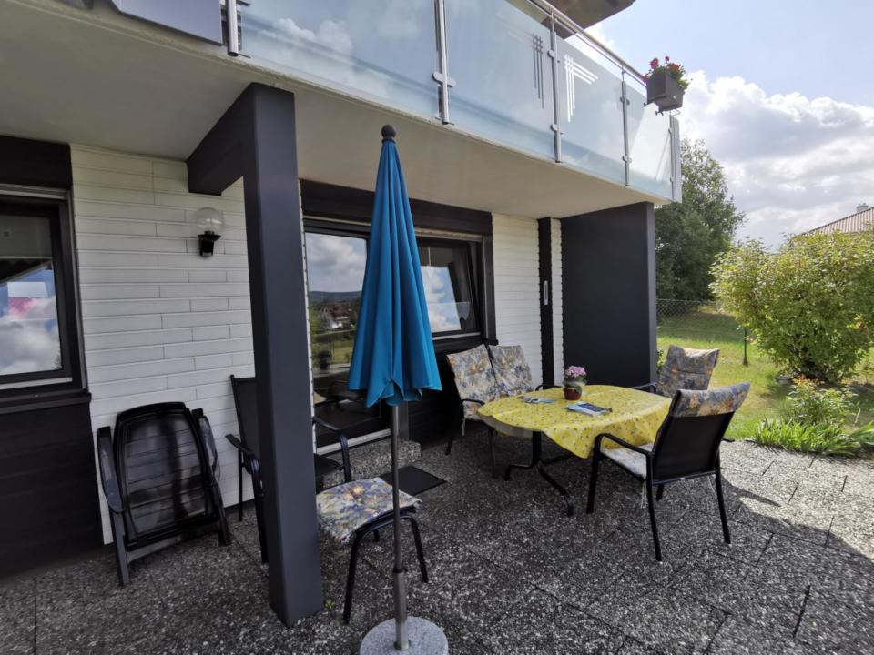 Eine Terrasse mit einem Tisch, auf dem eine gelbe Tischdecke liegt, vier Stühlen, einem blauen, geschlossenen Sonnenschirm und Topfpflanzen. Der Bereich befindet sich neben einem weißen Backsteinhaus mit großen Fenstern und einem verglasten Balkon darüber.