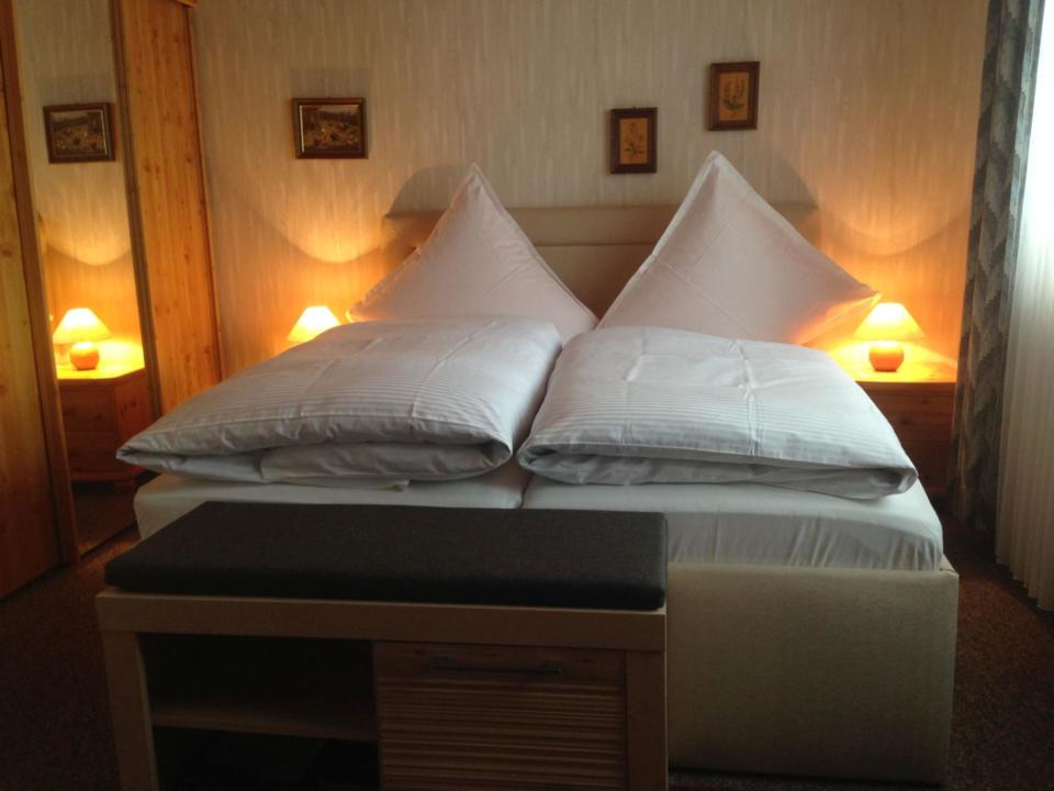 Ein ordentlich gemachtes Doppelbett mit weißer Bettwäsche, dreieckigen Kissen und Nachttischen mit brennenden Lampen. Über dem Bett hängen drei kleine Bilder, und am Fußende des Bettes steht eine Bank.