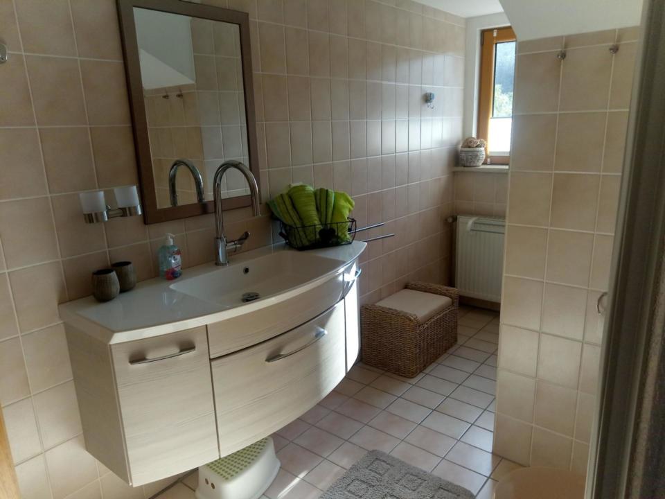 Ein Badezimmer mit beigen Fliesen, einem weißen Waschbecken und einem weißen Unterschrank, einem großen Spiegel, zwei Wasserhähnen, einem Weidenkorb mit grünen Handtüchern, einem Fenster, einem Heizkörper und einem kleinen Tritt auf dem Fliesenboden.