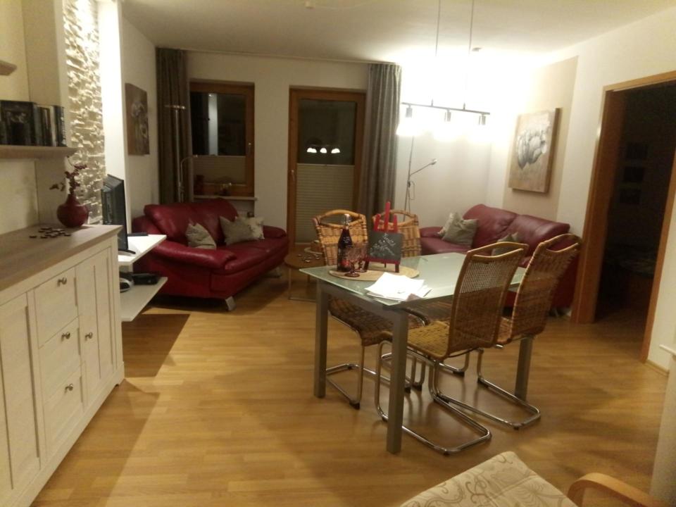 Ein gemütliches Wohnzimmer mit roten Sofas, einem gläsernen Esstisch mit vier Stühlen, Holzboden und Wandbildern. Auf dem Tisch stehen eine Flasche, Körbe und eine Geschenktüte, die eine warme, einladende Atmosphäre schaffen.