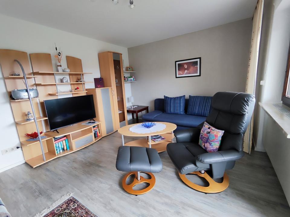 Ein gemütliches Wohnzimmer mit einem schwarzen Liegesessel, einem Fußhocker, einem blauen Sofa, Holzregalen, einem Fernseher, Büchern und Dekoartikeln. An einer grauen Wand neben einem Fenster mit cremefarbenen Vorhängen hängt ein gerahmtes Bild. Vor dem Sofa steht ein runder Couchtisch.