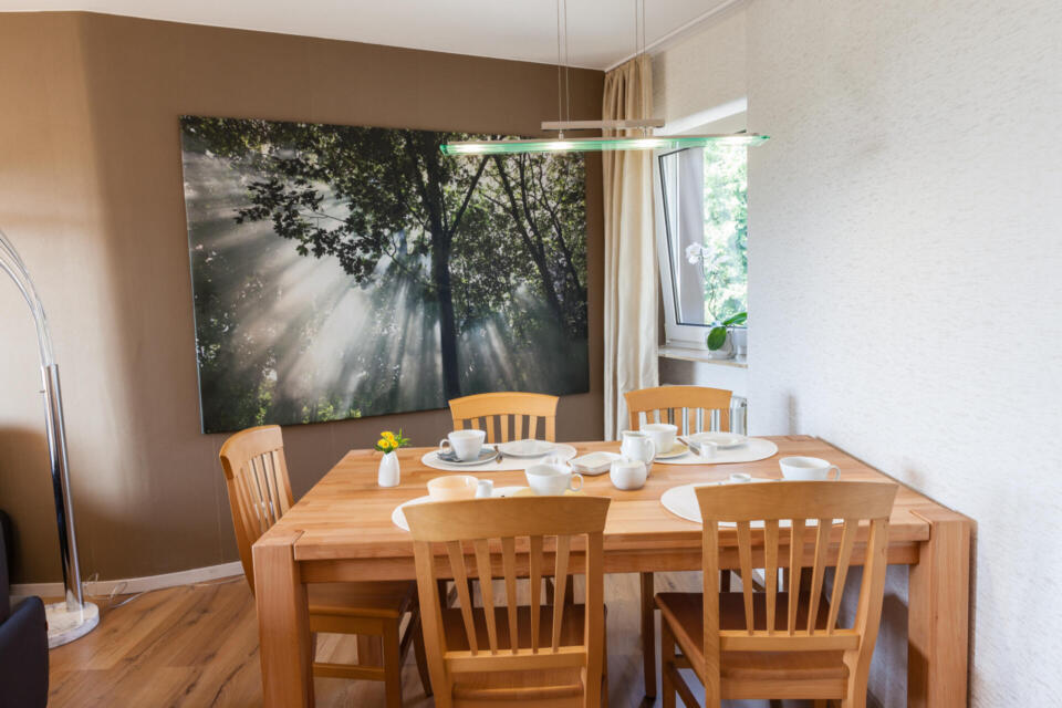 Ein Esszimmer mit einem Holztisch für sechs Personen, Holzstühlen, einem großen Wandbild mit sonnenbeschienenen Bäumen, einem Fenster mit Vorhängen und einer modernen Hängeleuchte über dem Tisch.