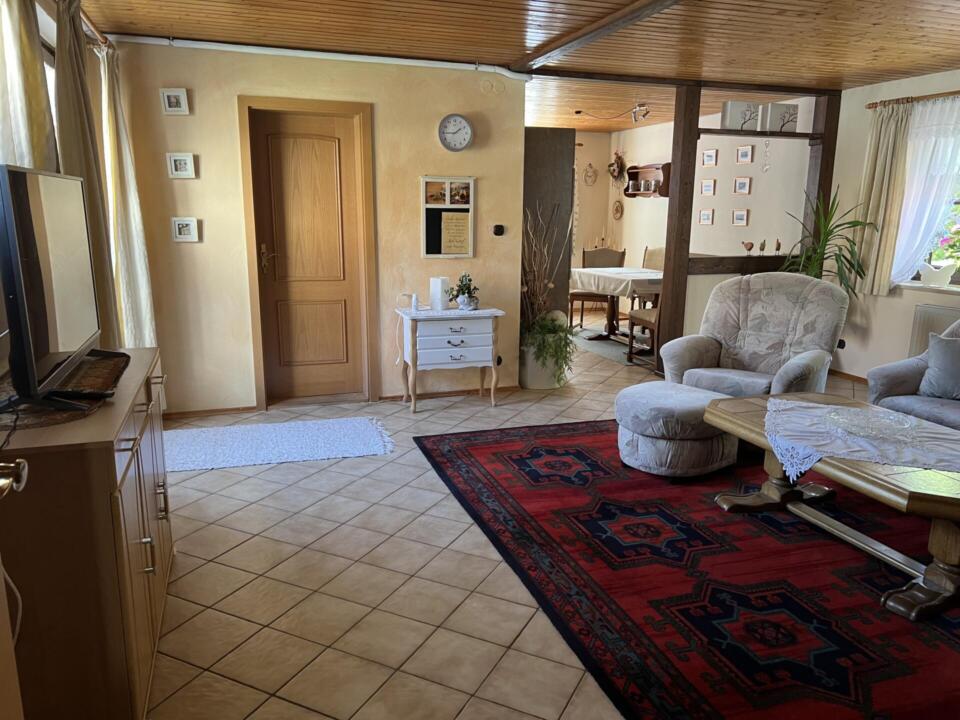 Ein gemütliches Wohnzimmer mit Fliesenboden, einem gemusterten roten Teppich, beigen Sesseln, einem Couchtisch aus Holz, einer Anrichte und einem Fernseher. Hellgelbe Wände, gerahmte Fotos, Pflanzen und eine Uhr schmücken den Raum. Durch die Fenster fällt Licht herein.