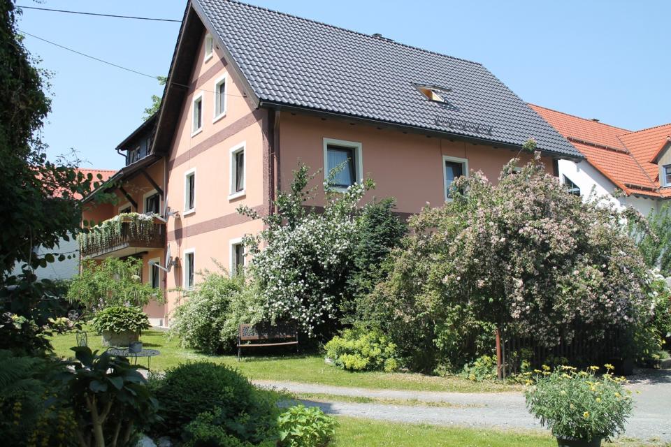 Ein zweistöckiges, pfirsichfarbenes Haus mit Satteldach, weiß umrahmten Fenstern und einem Balkon, umgeben von üppig grünen Sträuchern, blühenden Pflanzen und einem gepflegten Garten unter strahlend blauem Himmel.