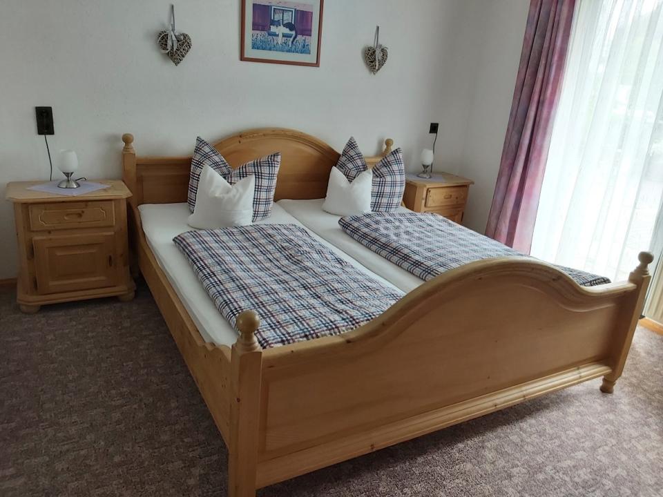 Ein gemütliches Schlafzimmer mit einem Doppelbett aus Holz, karierter Bettwäsche, zwei Nachttischen mit Lampen, herzförmigen Wanddekorationen und Vorhängen, die ein großes Fenster verdecken.