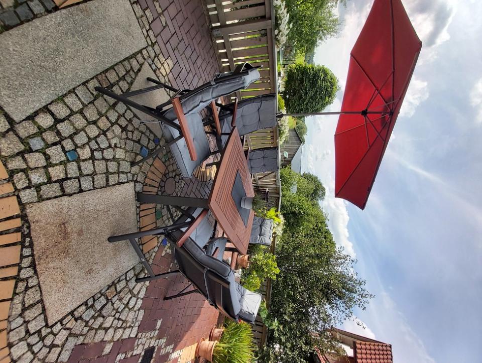 Eine Terrasse im Freien mit einem Holztisch, acht gepolsterten Stühlen, einem großen roten Sonnenschirm, Topfpflanzen und einer malerischen grünen Landschaft unter einem teilweise bewölkten Himmel. Der Bodenbelag aus Ziegeln und Stein verleiht der Kulisse zusätzlichen Charme.