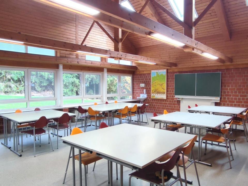 Ein Klassenzimmer im Schullandheim Weißenstadt mit Holzbalken, großen Fenstern, weißen Tischen, bunten Stühlen, einer Tafel und einer Wandkarte. Der Raum ist leer und wird durch Tageslicht aus Oberlichtern und Fenstern mit Blick ins Grüne hell beleuchtet.