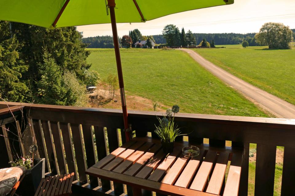 Ein Holzbalkon mit einem Tisch und einer Pflanze unter einem grünen Sonnenschirm bietet einen Blick auf eine ländliche Landschaft mit Feldern, Bäumen, einem Feldweg und Häusern in der Ferne unter einem klaren Himmel.