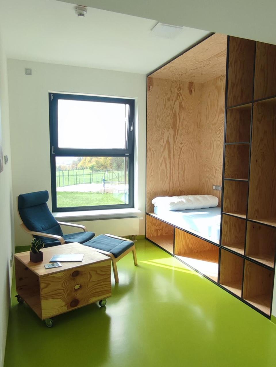 Ein modernes, minimalistisch eingerichtetes Zimmer im Schullandheim Weißenstadt verfügt über eine Schlafecke aus Holz, einen blauen Sessel mit Fußstütze, einen kleinen Rolltisch und eine Topfpflanze. Durch ein großes Fenster blickt man auf Wiesen und Bäume, während der Boden in einem leuchtenden Grün gehalten ist.