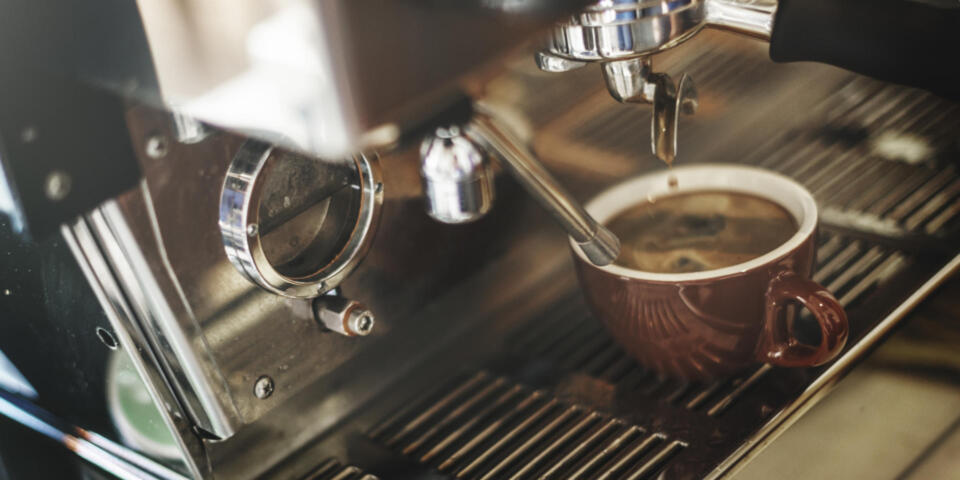 Nahaufnahme einer braunen Tasse, die aus einer Espressomaschine mit Kaffee befüllt wird. Die Dampfdüse und der Siebträger sind zu sehen, und der frisch gebrühte Kaffee bildet eine Crema auf der Oberfläche. Die Szene strahlt eine warme, einladende Atmosphäre aus.