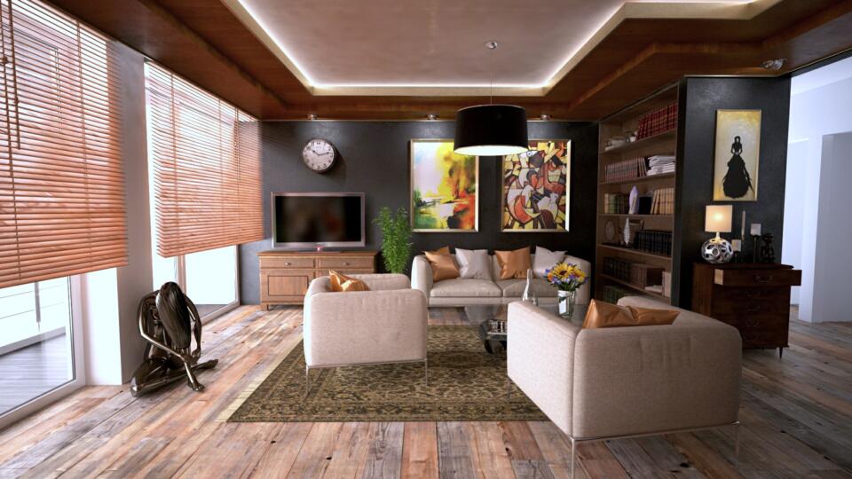 Modernes Wohnzimmer mit Holzböden, beigen Sofas, einem Fernseher, großen Fenstern mit Jalousien, farbenfrohen Kunstwerken an den Wänden, Bücherregalen, einem Teppich und einer schwarzen Pendelleuchte. Dekoartikel und Pflanzen runden das gemütliche Ambiente ab.