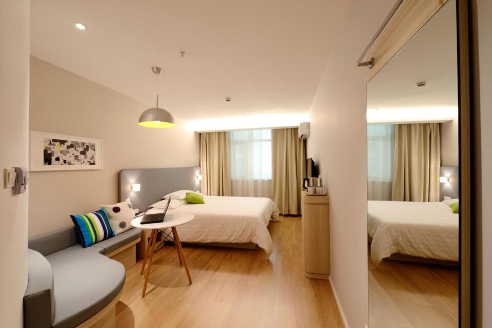 Modernes Hotelzimmer mit Holzböden, einem Doppelbett, einem Sofa, einem runden Tisch, einem großen Spiegel und einem Fenster mit beigen Vorhängen. Neutrale Farbtöne und eine schlichte Einrichtung sorgen für eine gemütliche, einladende Atmosphäre.