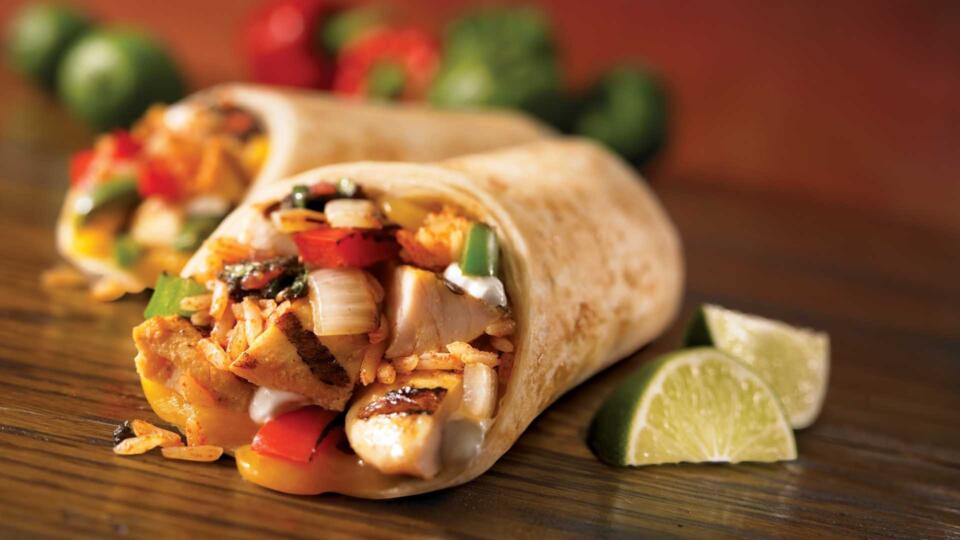 Zwei Burritos, gefüllt mit gegrilltem Hähnchenfleisch, Reis, Paprika, Zwiebeln und Soße, liegen auf einer Holzunterlage, im Hintergrund sind Limettenschnitze und verschwommene grüne Gemüsestücke zu sehen.