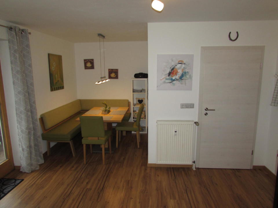 Essbereich mit einer grünen Eckbank, vier Stühlen, einem Holztisch, Wanddekoration, einer weißen Tür, einem Heizkörper und einem Holzboden. Links befindet sich ein Fenster mit Vorhängen, und über dem Tisch hängen zwei Pendelleuchten.