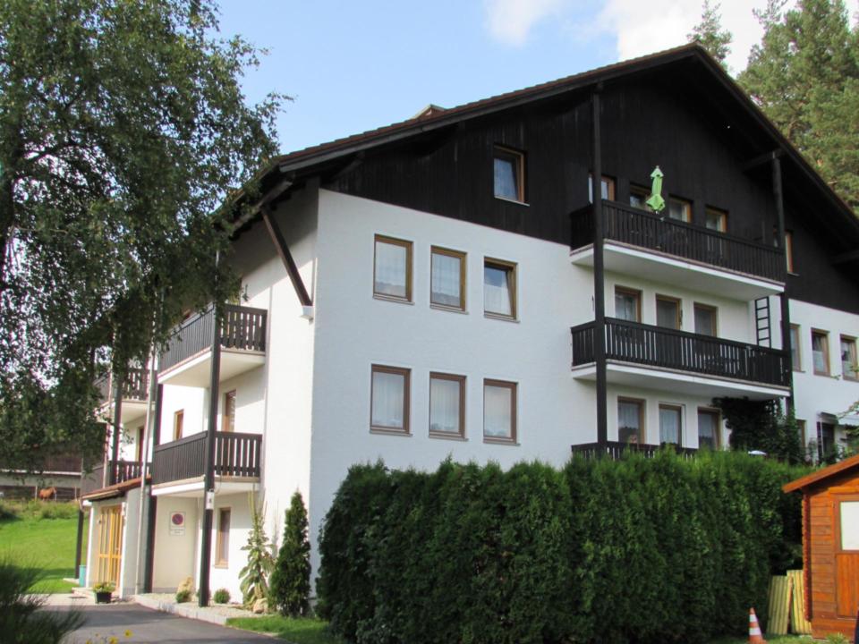 Weißes mehrstöckiges Gebäude mit dunklen Holzbalkonen und einem Satteldach, umgeben von grünen Sträuchern, Bäumen und Rasenflächen, in einer ruhigen, sonnigen Umgebung im Freien gelegen.