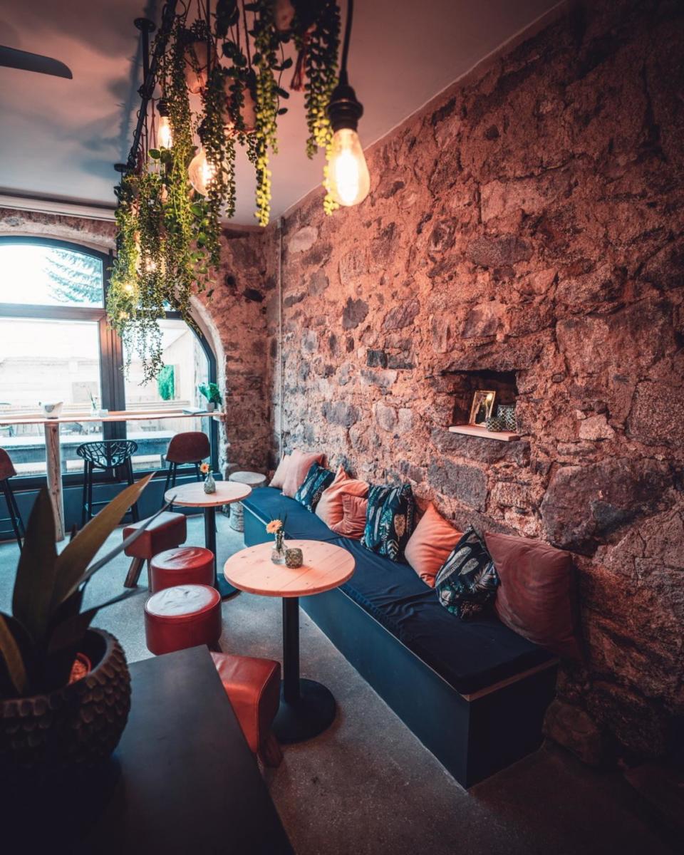 Gemütliches Café mit Steinwänden, hängenden Pflanzen und warmer Beleuchtung. An einer Seite steht eine lange, gepolsterte Bank mit Kissen, daneben kleine runde Tische und Stühle. Durch das große Fenster fällt Tageslicht herein.