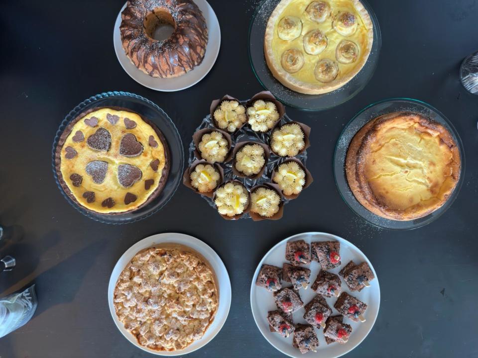 Auf einem schwarzen Tisch sind sieben verschiedene gebackene Desserts ausgestellt, darunter Kuchen, Muffins, Törtchen und Brownies, die von oben betrachtet kreisförmig angeordnet sind.