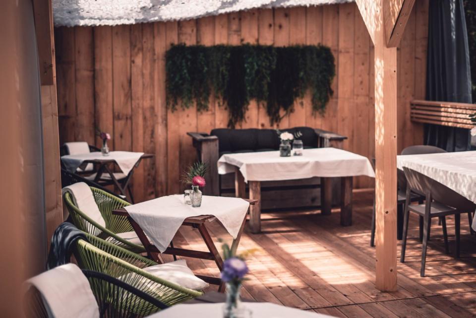Gemütlicher Essbereich im Freien mit Holzwänden, mehreren Tischen mit weißen Tischdecken, Topfpflanzen als Tischdekoration, grünen Stühlen und einfallendem Sonnenlicht. Im Hintergrund ziert Grün die Wand.