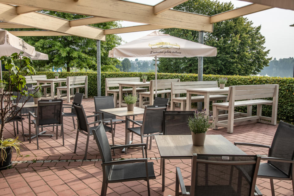 Eine Terrasse im Freien mit schwarzen Stühlen, weißen Holzbänken und Tischen mit kleinen Topfpflanzen. Über den Köpfen stehen weiße Sonnenschirme, und im Hintergrund sind grüne Bäume zu sehen, was eine ruhige und einladende Atmosphäre schafft.