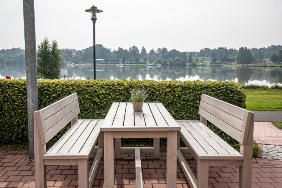 Auf einer mit Ziegeln gepflasterten Terrasse am See, die von grünen Hecken gesäumt ist, steht ein Holztisch mit zwei Bänken. Im Hintergrund sind an einem bewölkten Tag Bäume und ein Laternenpfahl zu sehen.