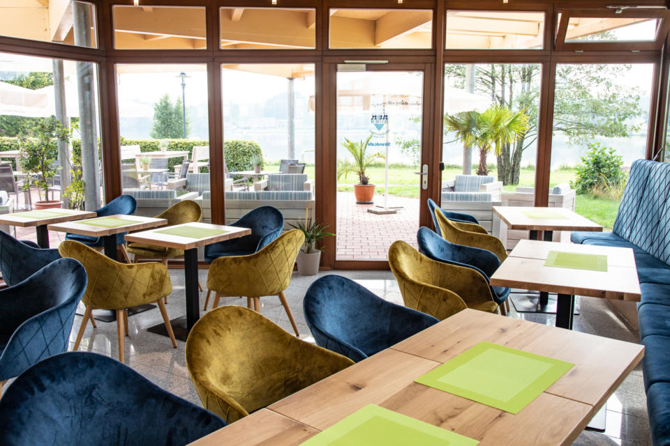 Ein modernes Café mit Holztischen, blauen und gelben Stühlen, grünen Tischsets, großen Fenstern und Blick auf eine Terrasse mit Sitzgelegenheiten im Freien, viel Grün und Palmen in der Nähe eines Gewässers.