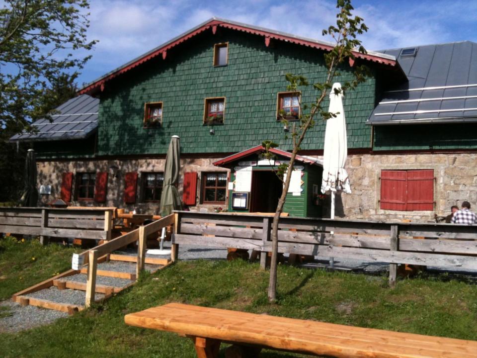 Ein rustikales, grünes Holzgebäude mit roten Fensterläden, Sitzgelegenheiten im Freien und Sonnenschirmen, umgeben von Bäumen und unter blauem Himmel. Im Vordergrund sind ein Holzzaun und Picknicktische zu sehen.