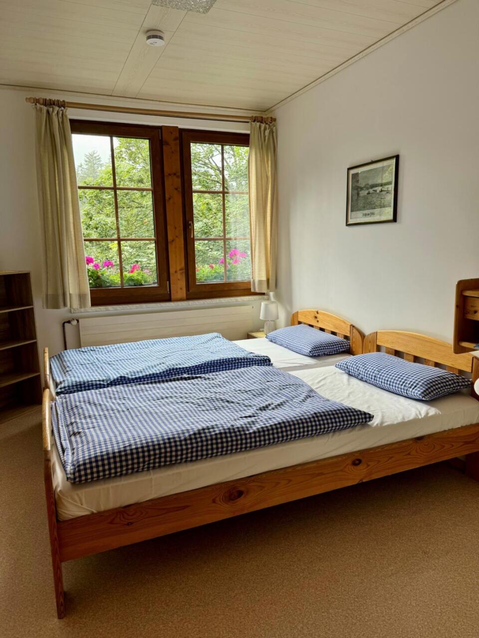 Ein gemütliches Schlafzimmer im Waldsteinhaus verfügt über ein Doppelbett mit blau karierter Bettwäsche, einen Holzrahmen und ein Fenster, das mit beigen Vorhängen geschmückt ist. An der Wand hängt ein gerahmtes Bild, und ein Regal sorgt für zusätzlichen Charme. Vom Fenster aus genießen Sie den Blick auf Bäume und Blumen – so haben Sie die Natur direkt vor der Haustür.
