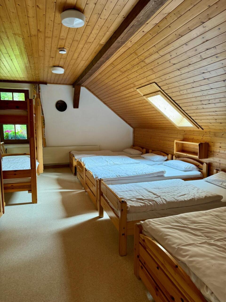 Ein gemütliches Schlafzimmer im Dachgeschoss des Waldsteinhauses verfügt über vier Einzelbetten aus Holz, weiße Bettwäsche, holzvertäfelte Wände und Decken, ein Dachfenster sowie ein Fenster, durch das Tageslicht hereinfällt.