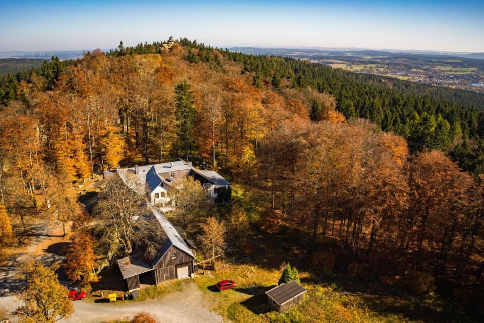 Luftaufnahme des Waldsteinhauses, umgeben von herbstlich gefärbten Bäumen an einem Hang; unter dem strahlend blauen Himmel sind ein Wald und eine Stadt in der Ferne zu sehen.