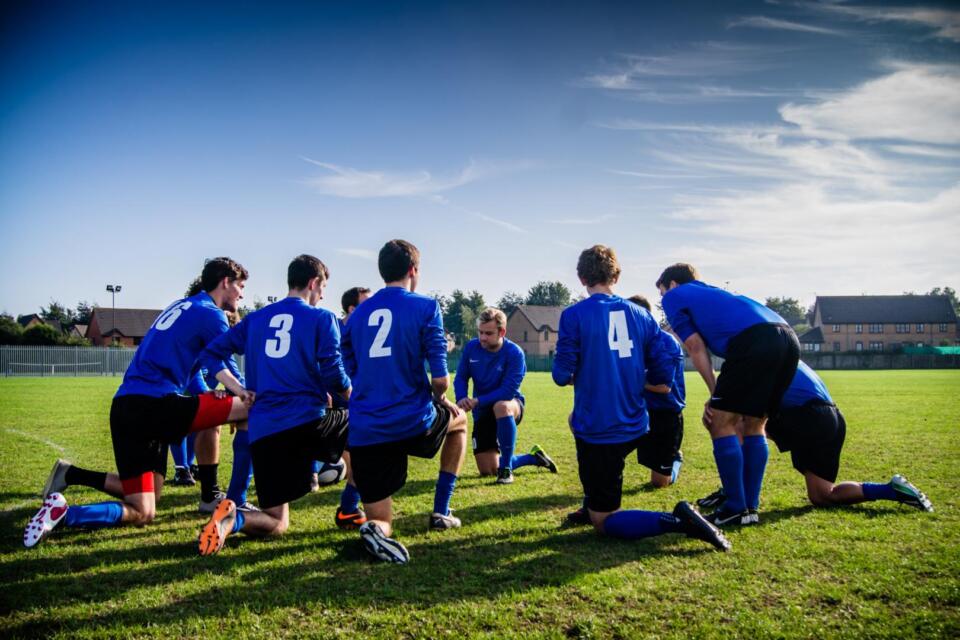 Eine Fußballmannschaft in blauen Trikots kniet auf dem Rasen und hört ihrem Trainer während eines Trainings oder Spiels zu, im Hintergrund sind Häuser und Bäume vor einem blauen Himmel zu sehen.