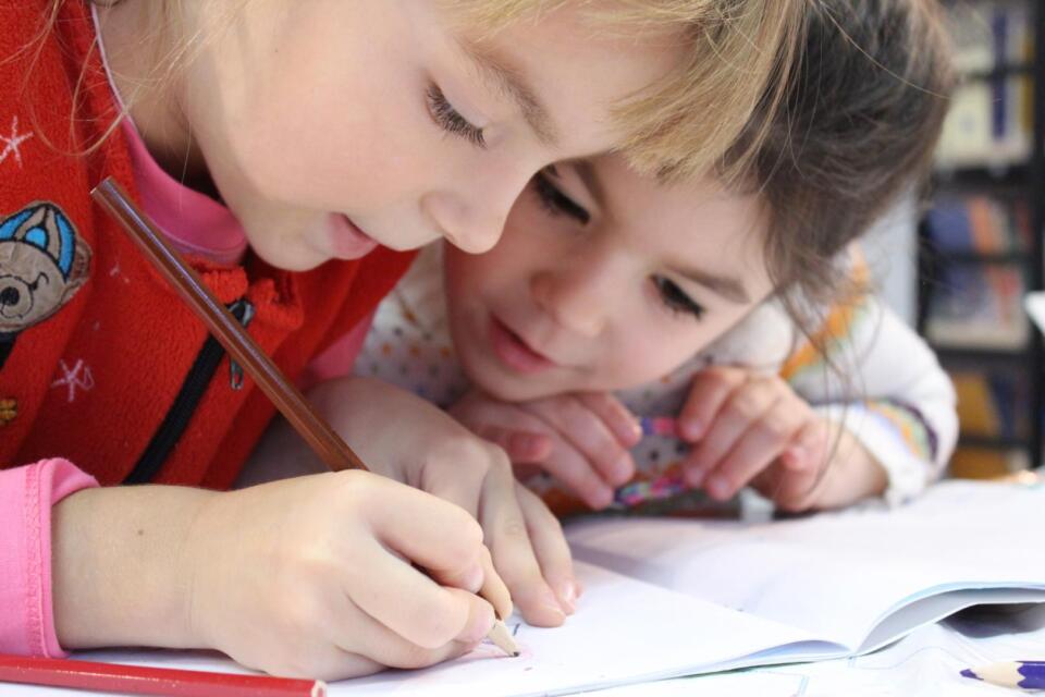Zwei kleine Mädchen zeichnen oder schreiben gemeinsam mit Buntstiften auf weißem Papier und sind bei einer kreativen Aktivität im Rahmen der Kinderbetreuung ganz in ihre Arbeit vertieft.