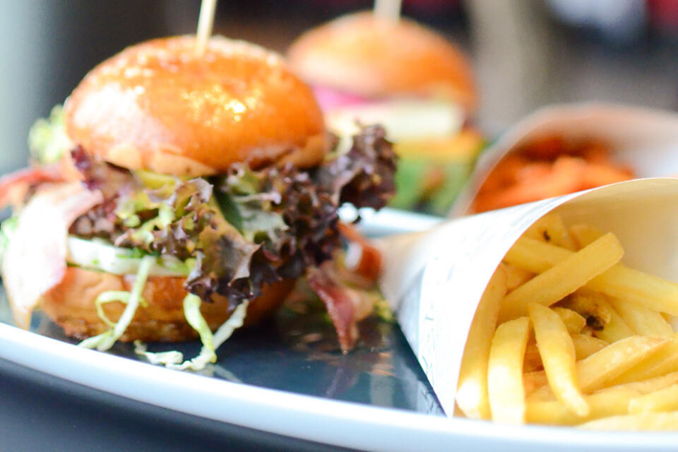 Im Bild ist eine Nahaufnahme eines Burgers mit Salat, Tomate und weiteren Belägen auf einem Brötchen zu sehen, daneben auf einem Teller eine mit Pommes frites gefüllte Papiertüte – eine appetitliche Szene, die an die herzhaften Gerichte erinnert, die im Steakhaus Siebenquell serviert werden.