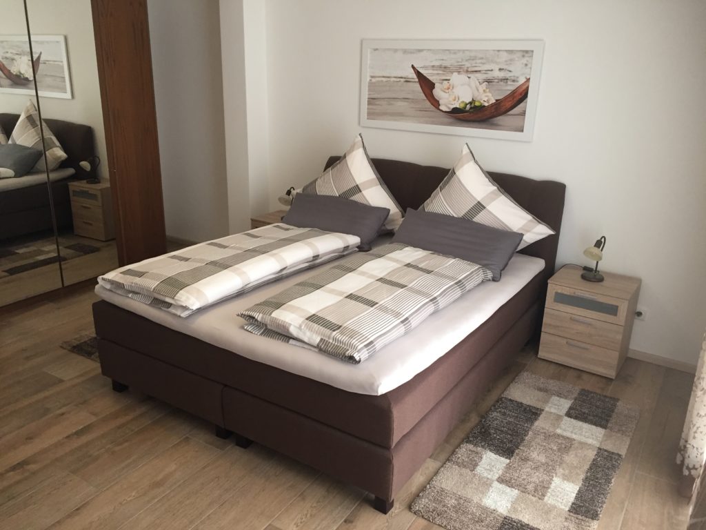In einem gemütlichen Schlafzimmer mit Holzboden und Teppichen steht ein ordentlich gemachtes Bett mit karierter Bettwäsche, zwei Kissen, zwei Nachttischen mit Lampen, einem Wandbild über dem Bett und einem Spiegelschrank.