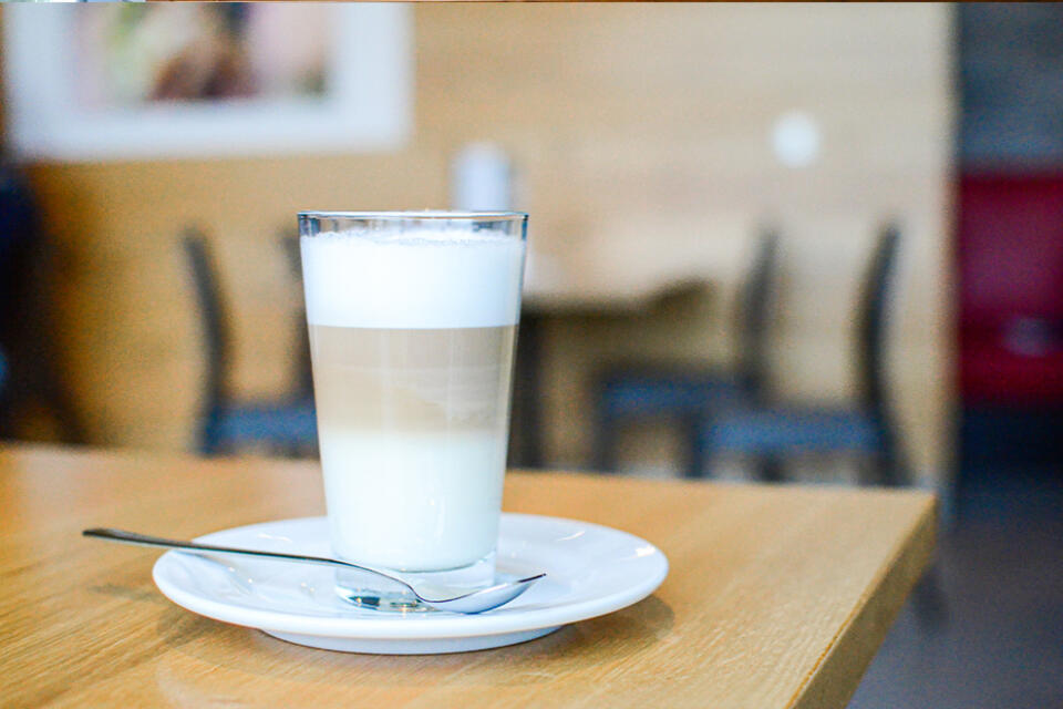 Ein Glas mit einem geschichteten Latte und Milchschaum steht auf einer weißen Untertasse mit einem Löffel darauf, platziert auf einem Holztisch in der sanft verschwommenen Café-Kulisse des Steakhauses Siebenquell.