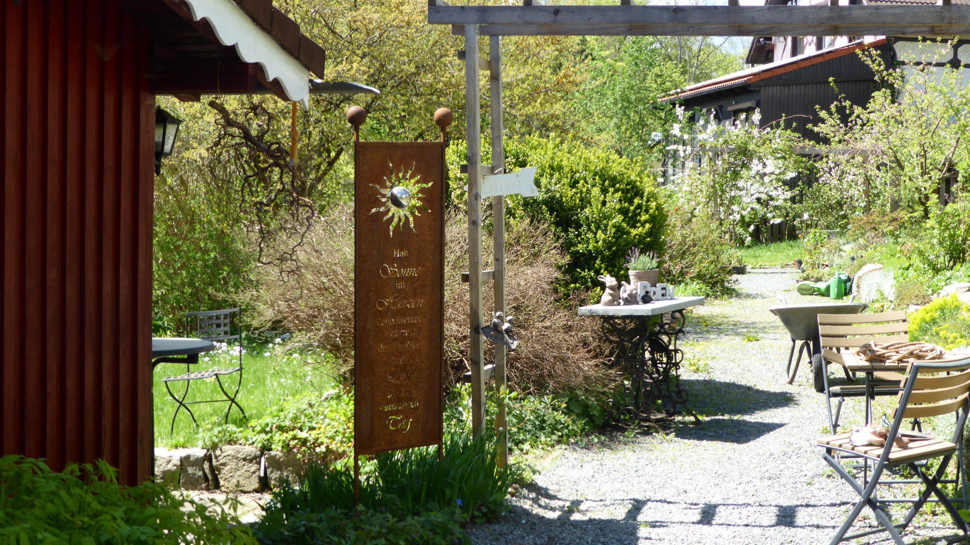 Im Lehr- und Sichtgarten befindet sich ein Gartencafé mit Tischen und Stühlen im Freien, einem dekorativen Metallschild und üppigem Grün. Sonnenlicht erhellt den Kiesweg, der zu einem rustikalen Gebäude im Hintergrund führt, und unterstreicht die ruhige Atmosphäre dieser malerischen Kulisse.